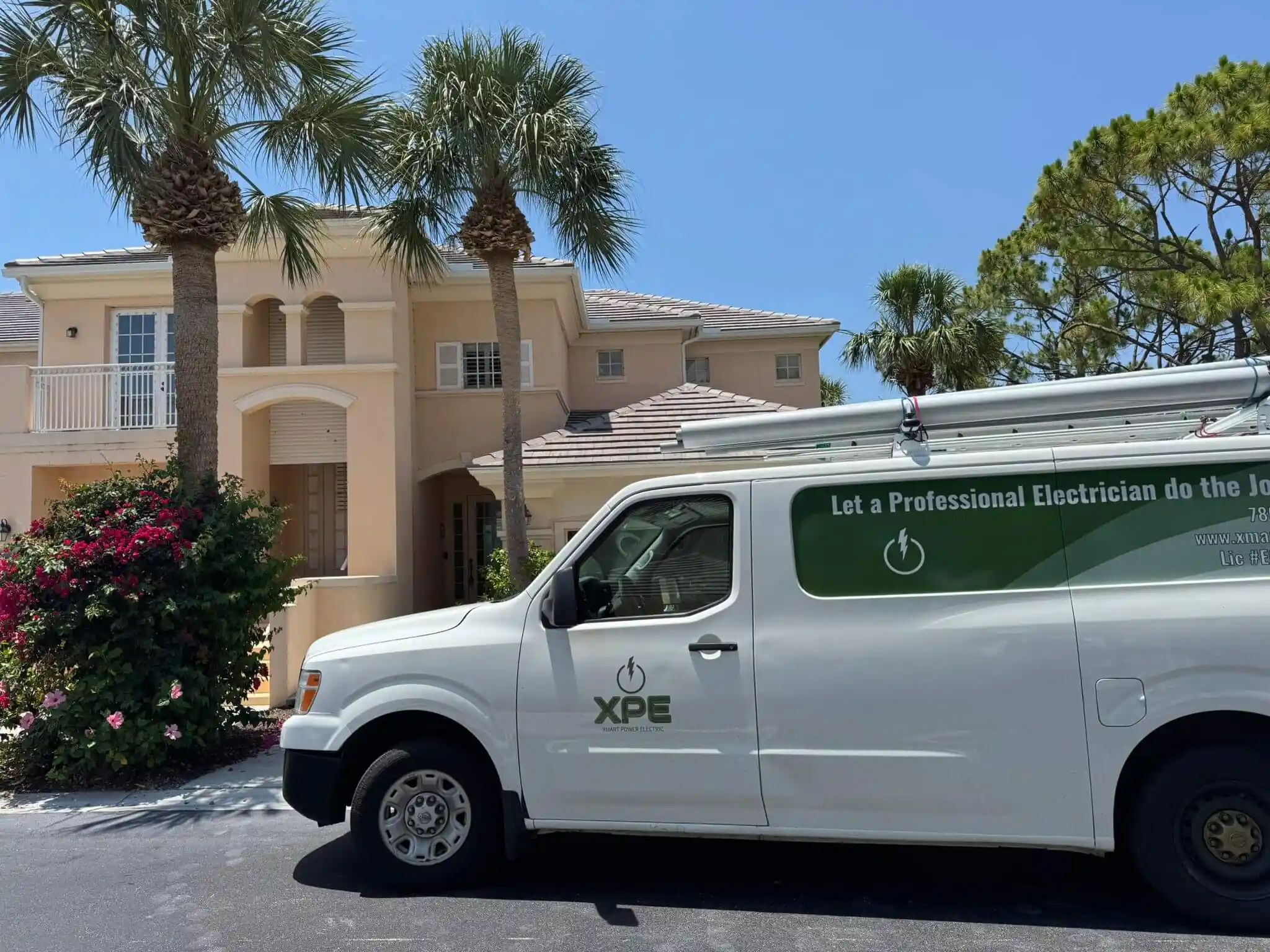 Service van from the electrical company XPE parked in front of a two-story residential house with palm trees and flowering bushes. The van has a sign that reads "Let a Professional Electrician do the Job."
