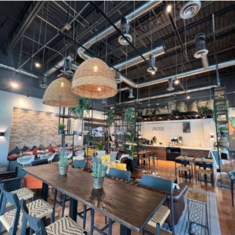 Cozy cafe interior with a large communal wood table, rattan pendant lights, exposed industrial ceiling, and a seating nook with a brick accent wall.