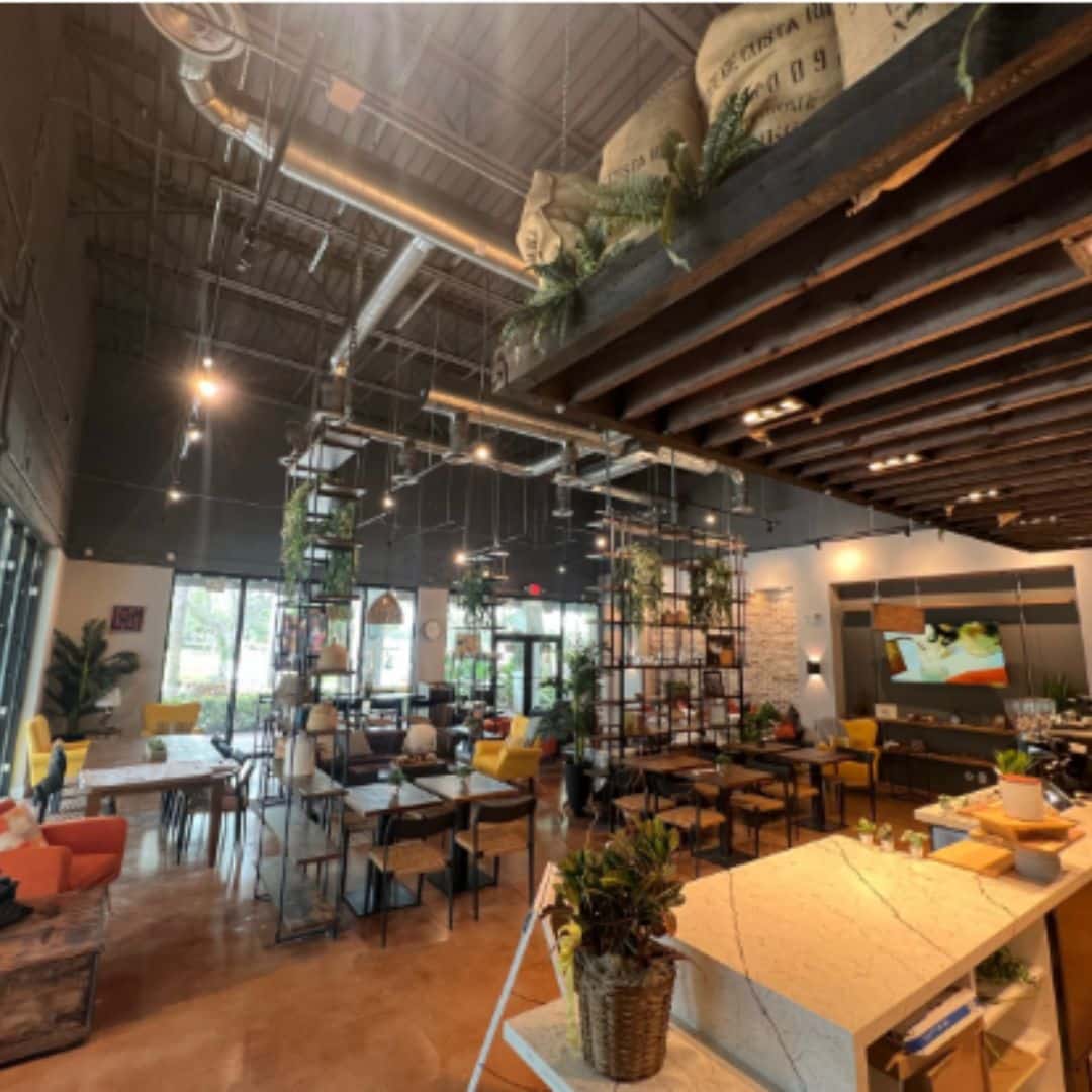 Industrial-style cafe or restaurant interior with high ceilings, exposed ductwork, dark structural shelving, orange concrete floor, and rustic seating areas.
