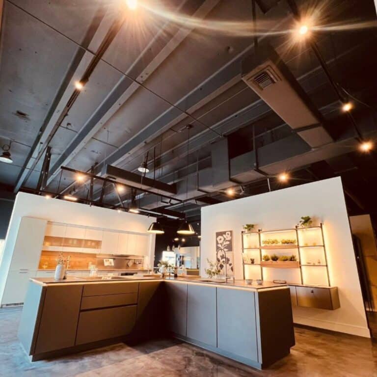 Modern kitchen showroom with dark, industrial ceilings and track lighting illuminating sleek gray and white cabinetry displays.