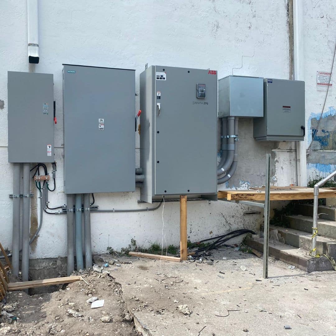 Exterior commercial electrical installation featuring a row of large gray utility and transfer switch panels from brands like Siemens and ABB mounted on a white wall.